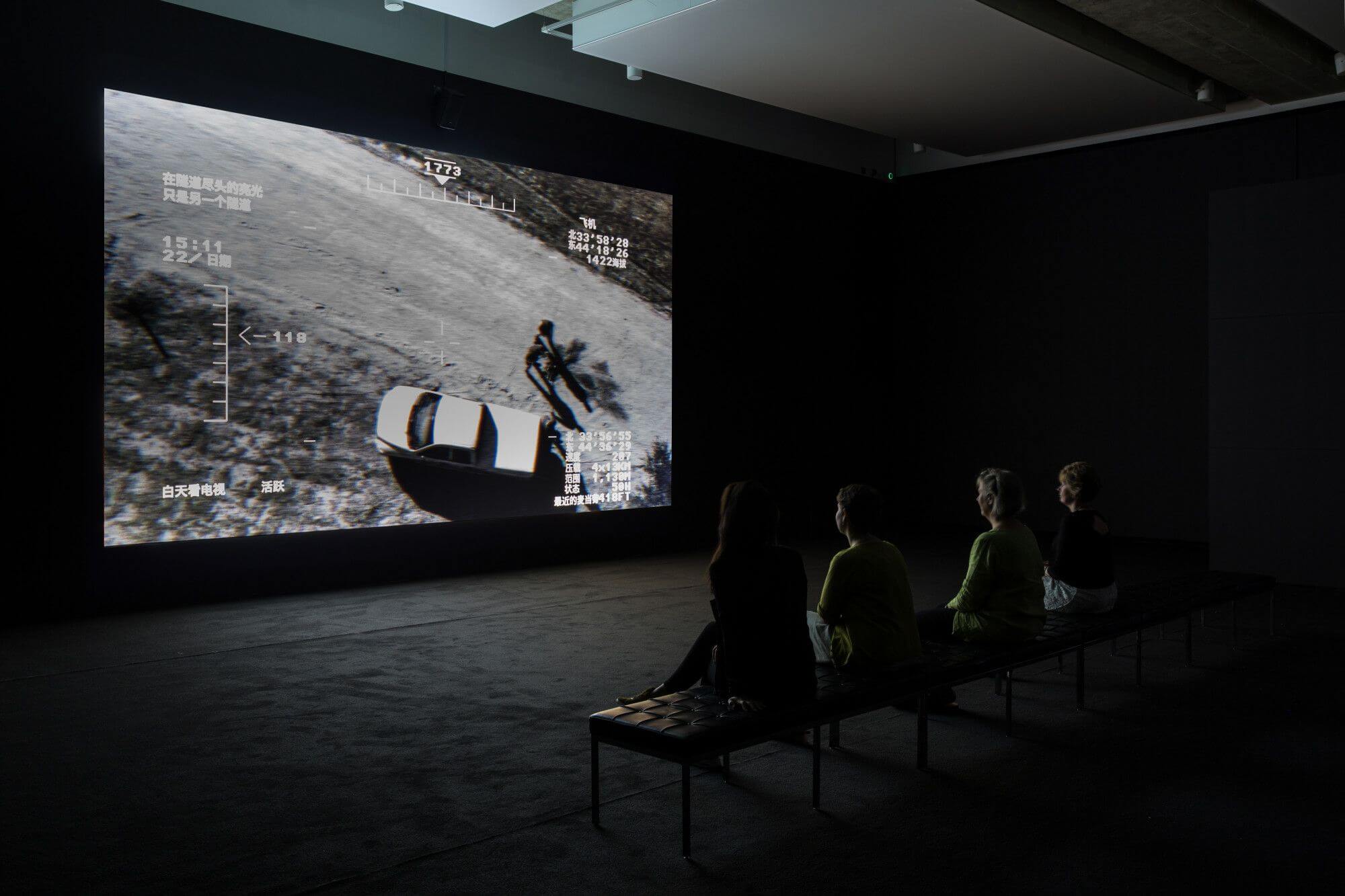 People sit on a bench in a dark gallery room, where a black and white film is screening